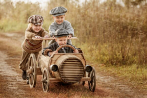 Two young children ride in the third race car from wooden barrels on a rural road autumn evening